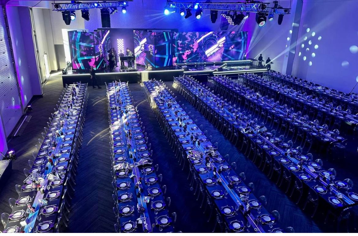 Large-scale corporate gala dinner with banquet tables, LED stage backdrop, dramatic blue and purple lighting, and professional event production setup