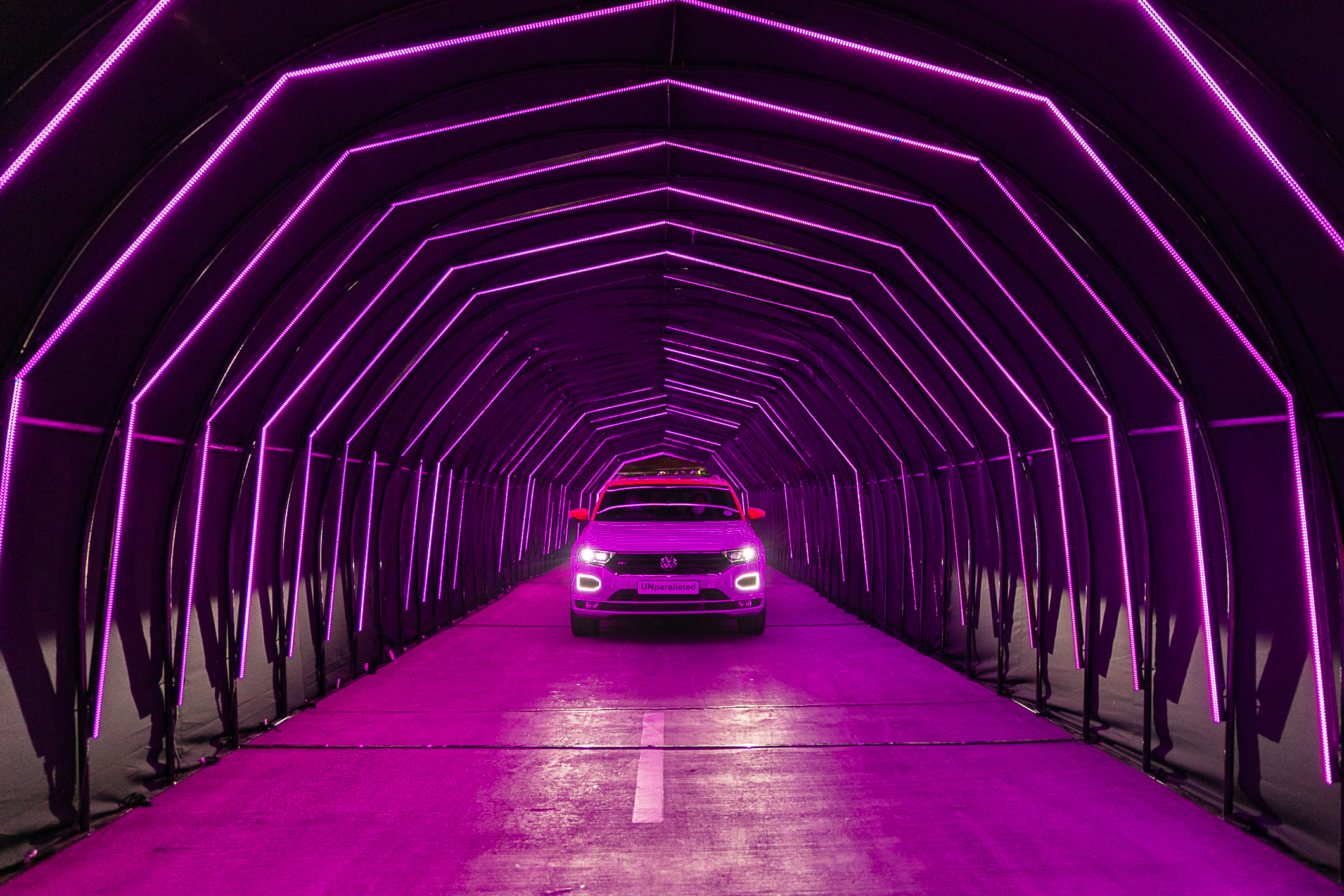 A Volkswagen SUV drives through a dramatic purple LED light tunnel during a theatrical automotive brand activation event, with the word "UNparalleled" displayed on the vehicle's number plate.