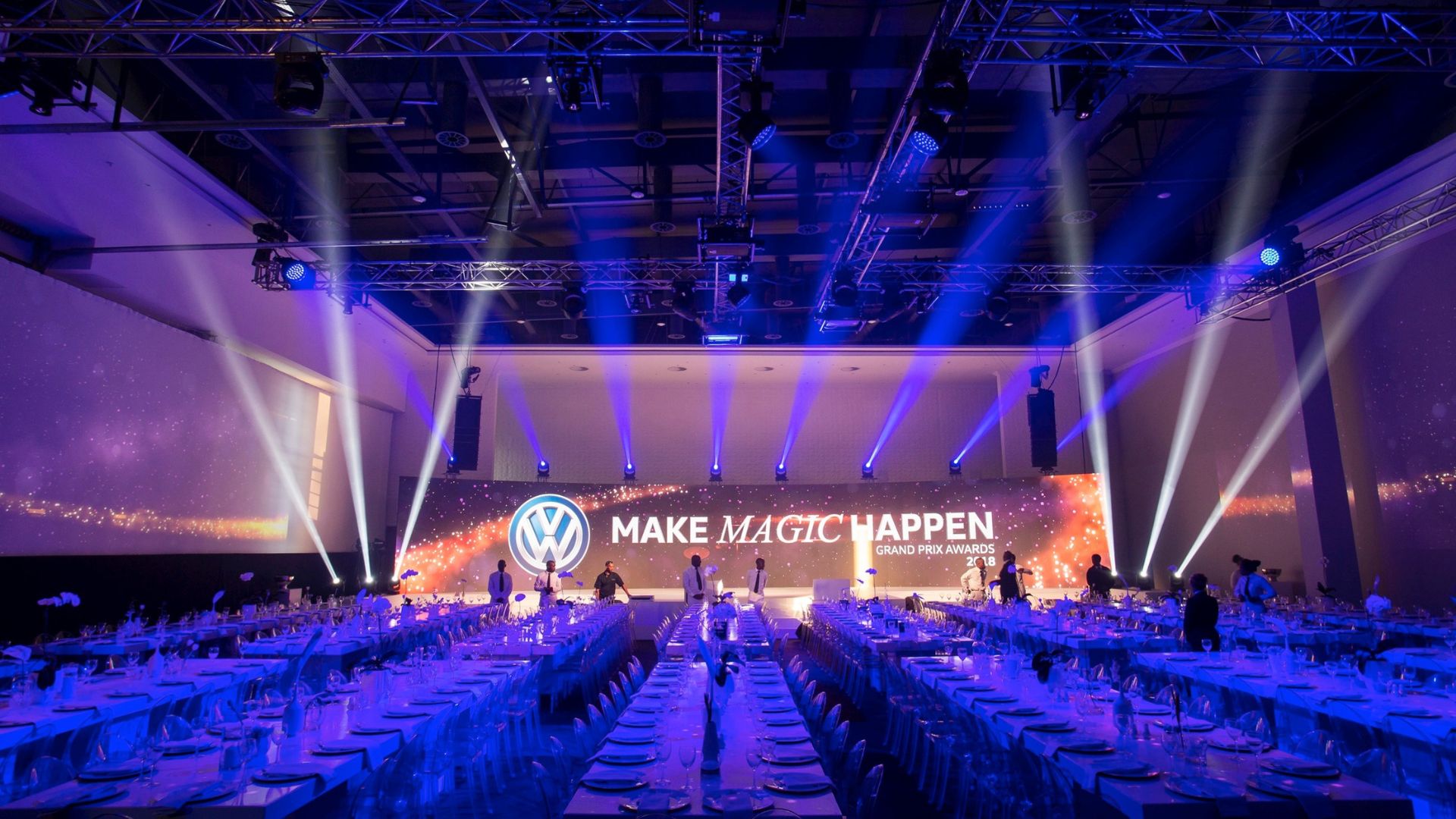 A large-scale corporate gala dinner setup produced by Mushroom Productions for the Volkswagen Grand Prix Awards 2018, featuring dramatic blue beam lighting, a branded LED backdrop reading "Make Magic Happen" and rows of elegantly set banquet tables.