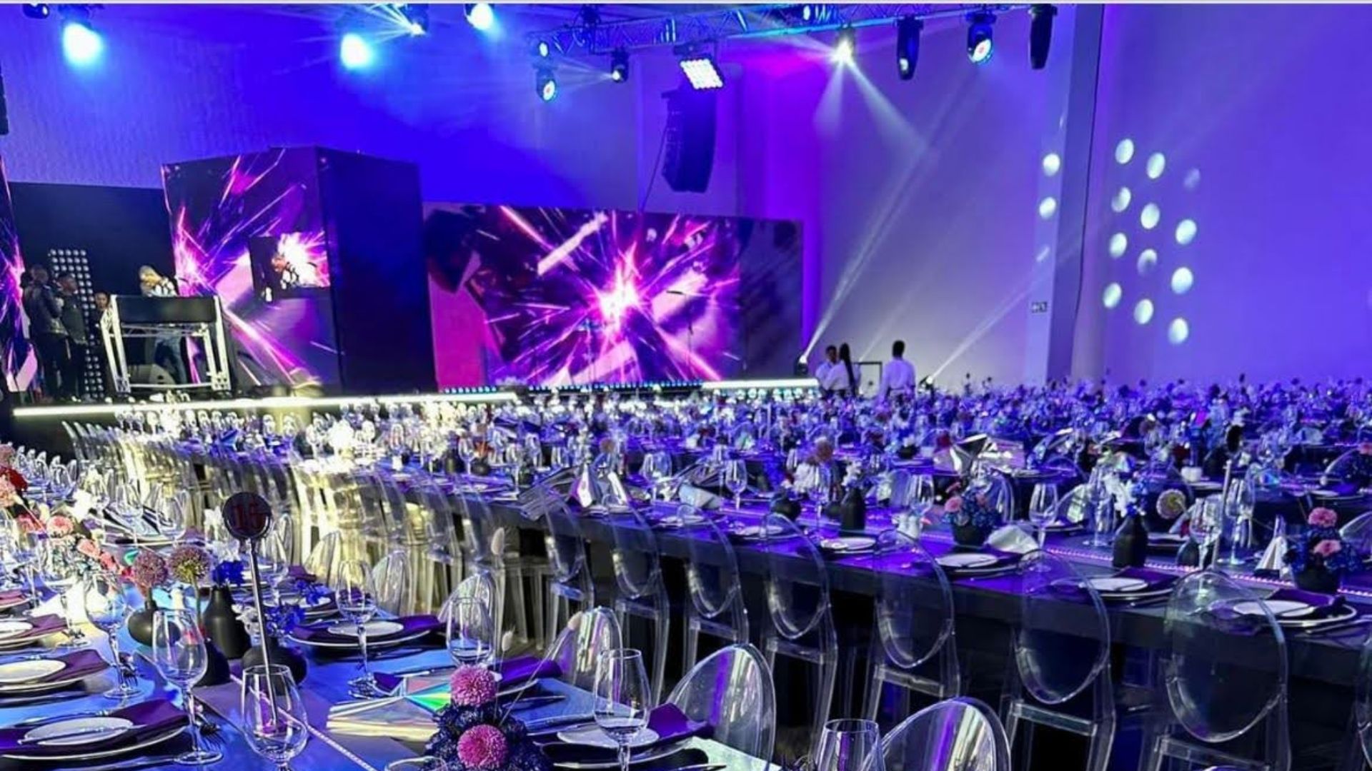 Mushroom Productions corporate gala dinner setup featuring long banquet tables with deep blue and purple linen, ghost chairs, and floral centrepieces, set against dramatic purple wash lighting, LED screens and full production staging in a large-scale South African event venue